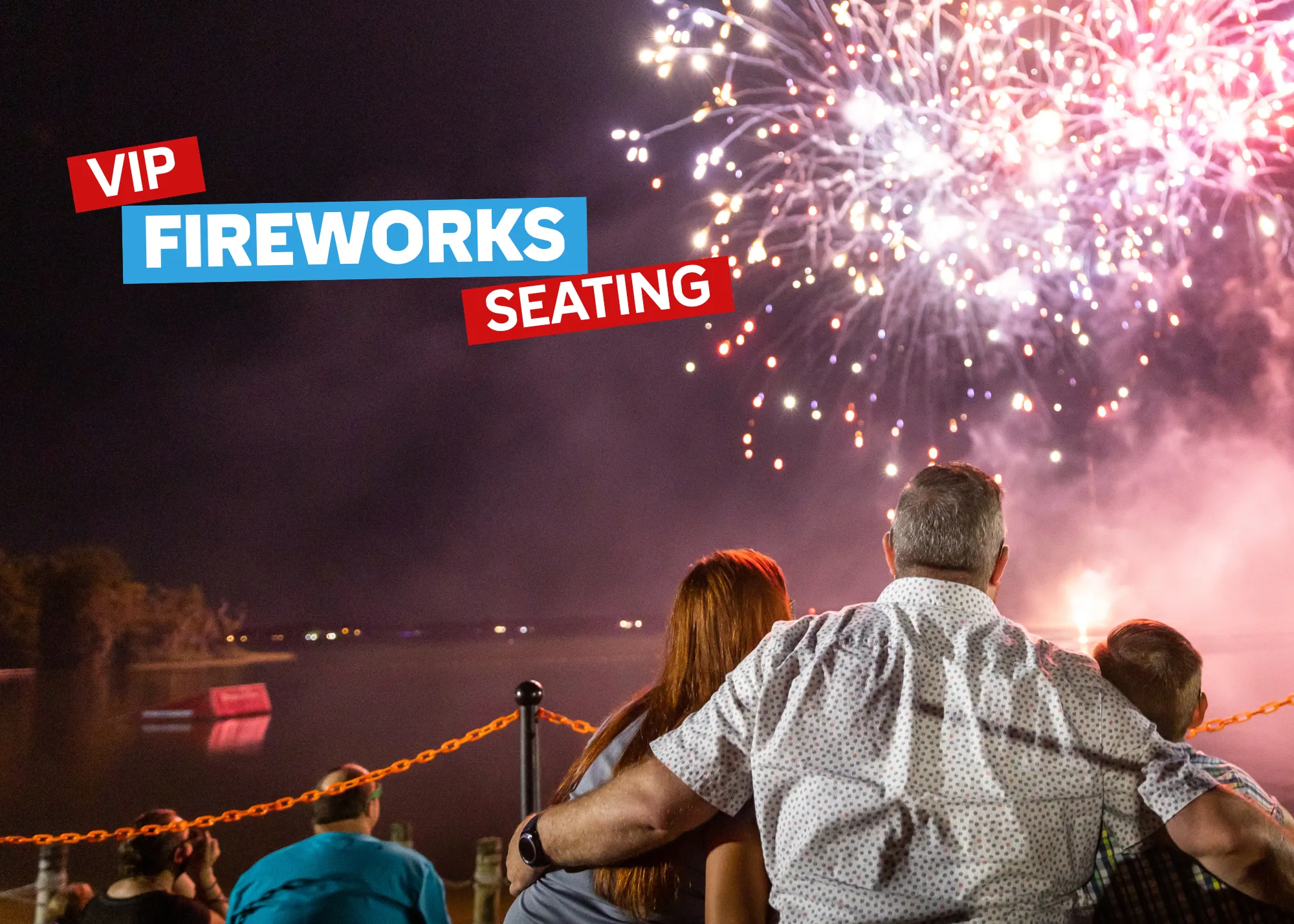 Family of three huddled together in a stadium at LEGOLAND® Florida Resort, smiling as colorful fireworks light up the night sky above.